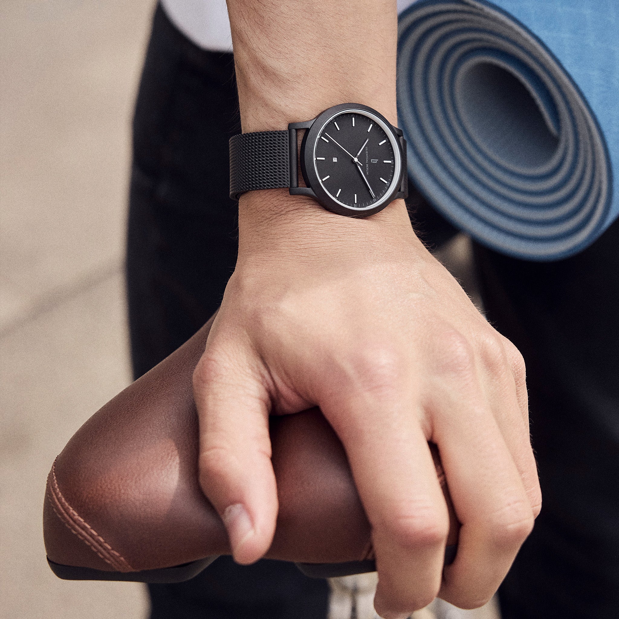 Minimalist black wristwatch with mesh strap on wrist, hand holding brown bicycle seat, yoga mat