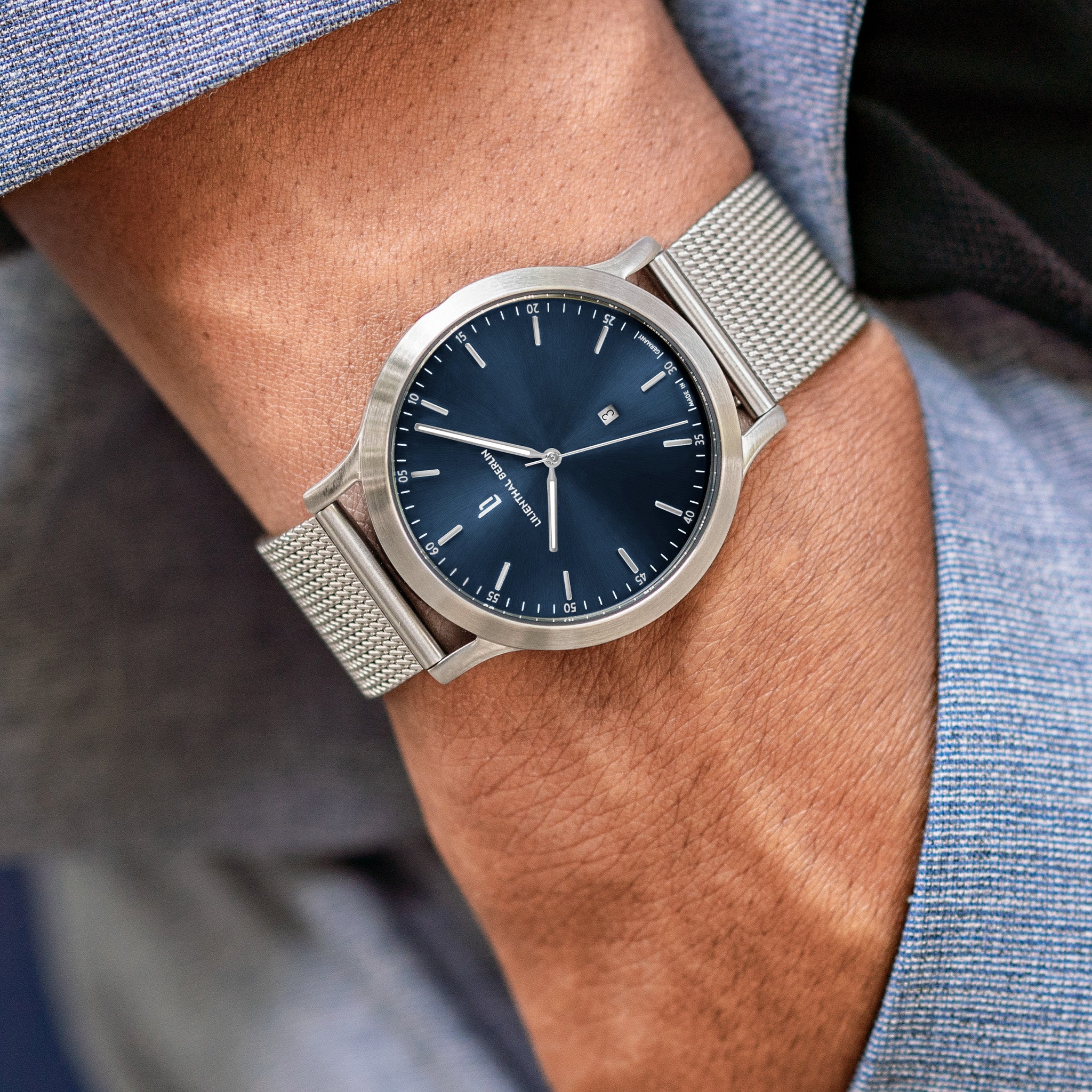 Minimalist silver mesh strap wristwatch with blue dial on wrist, worn with a blue suit