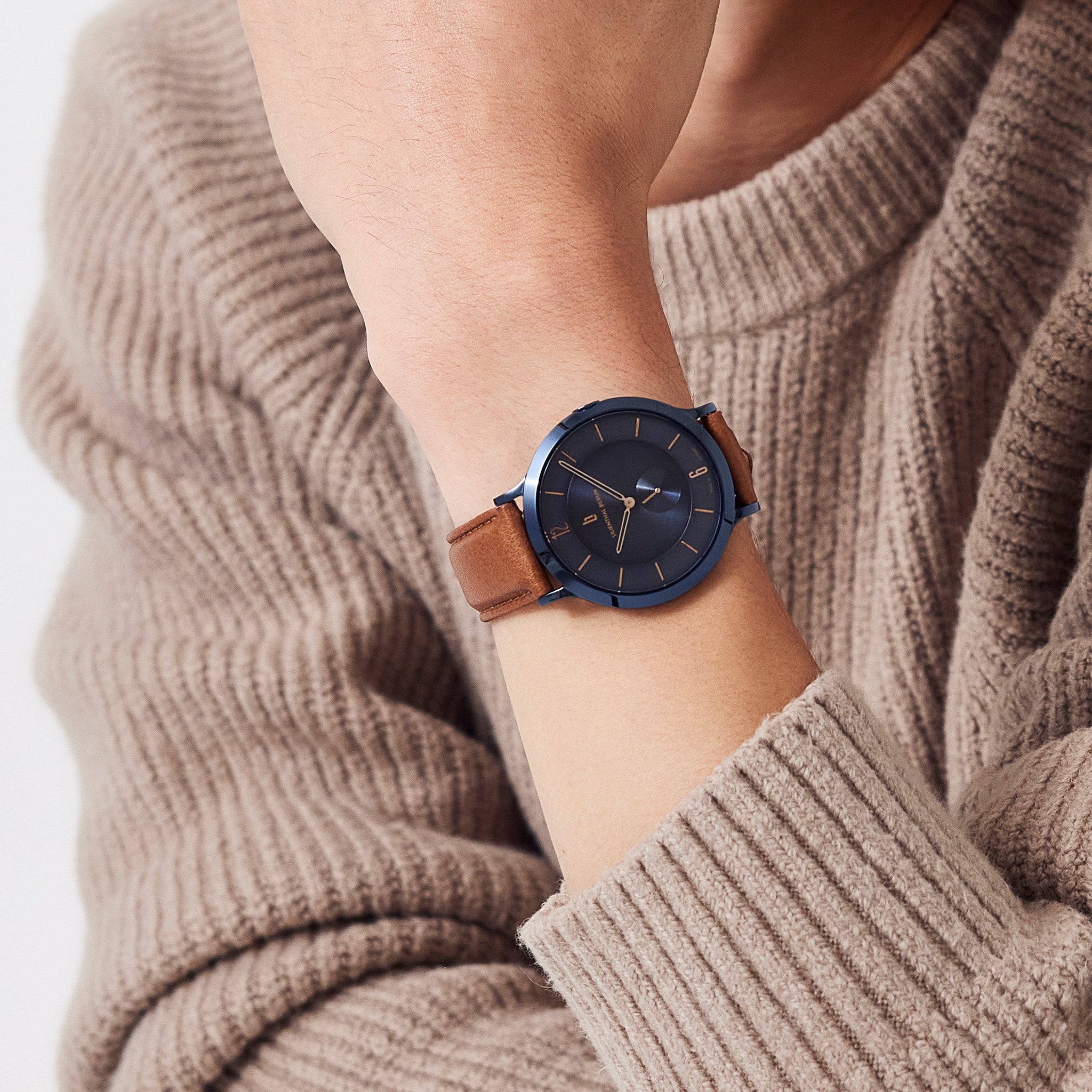 Minimalist blue watch with brown leather strap on wrist, worn over beige knit sweater.