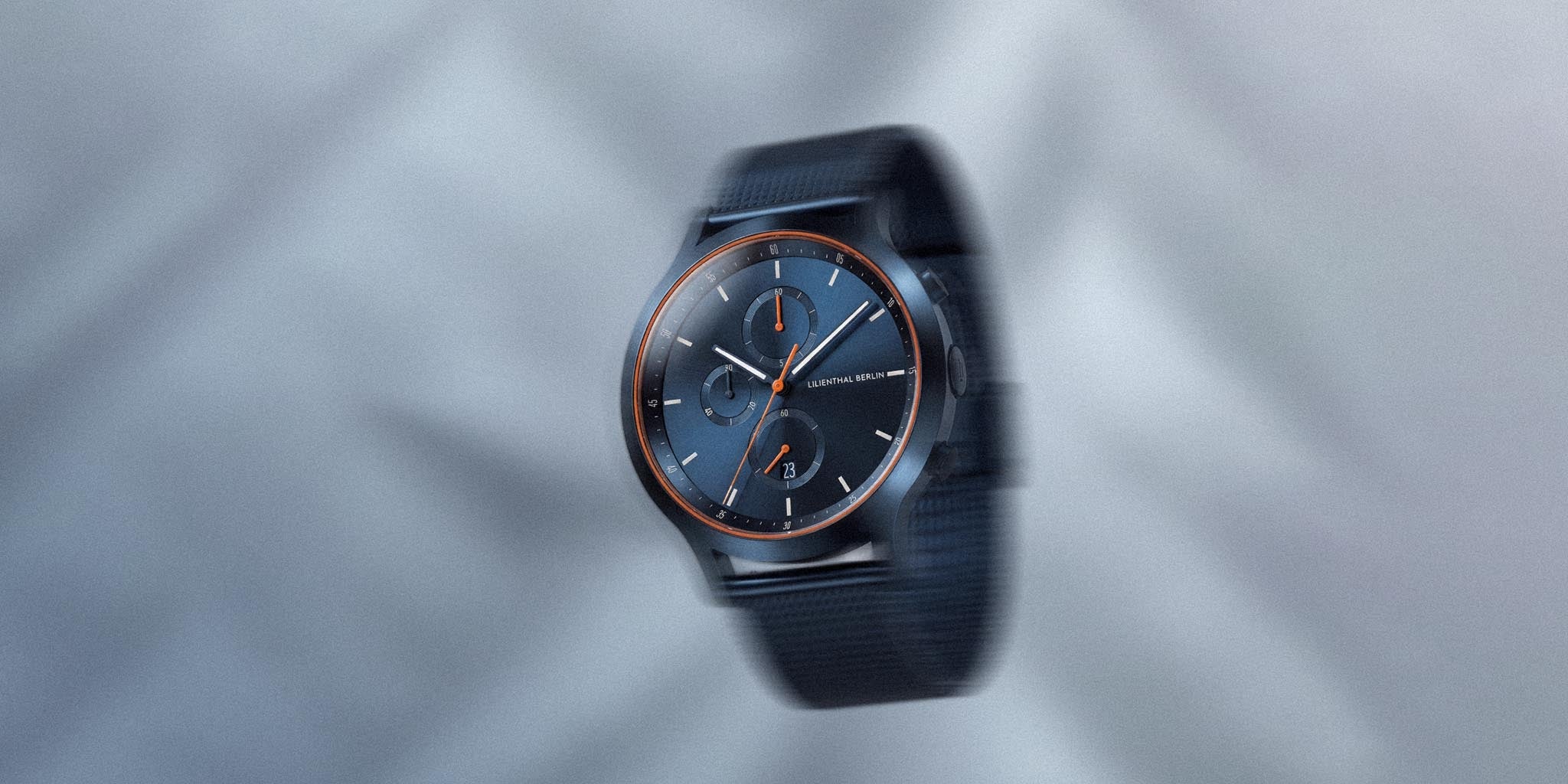 Blue chronograph watch with orange accents and mesh strap on a light background
