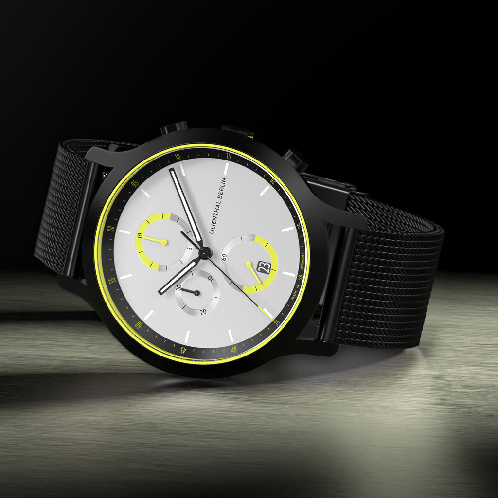 Modern chronograph watch with black mesh strap, silver dial, and yellow accents on a dark background.