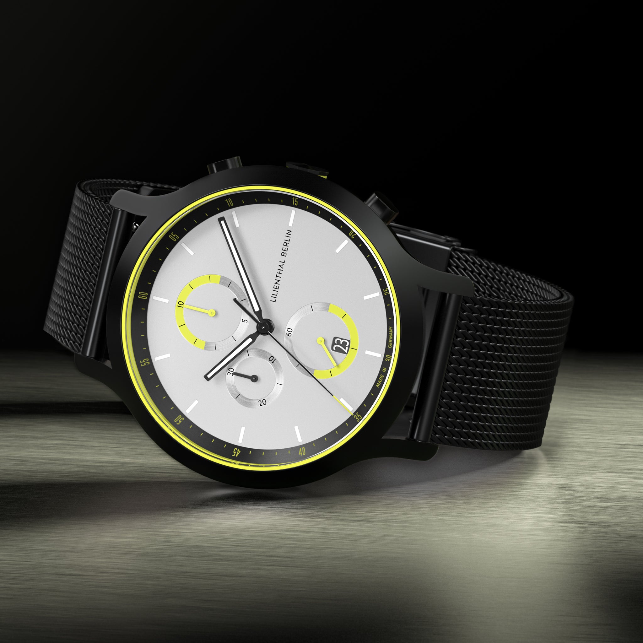 Modern chronograph watch with black mesh strap, silver dial, and yellow accents on a dark background.