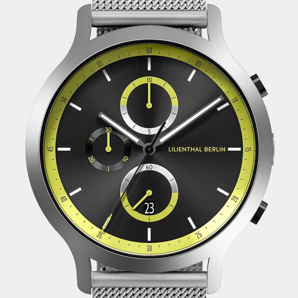 Modern chronograph watch with black dial, yellow accents, and stainless steel mesh band.