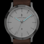 Minimalist silver watch with white dial, glowing blue hands, date display, brown leather strap