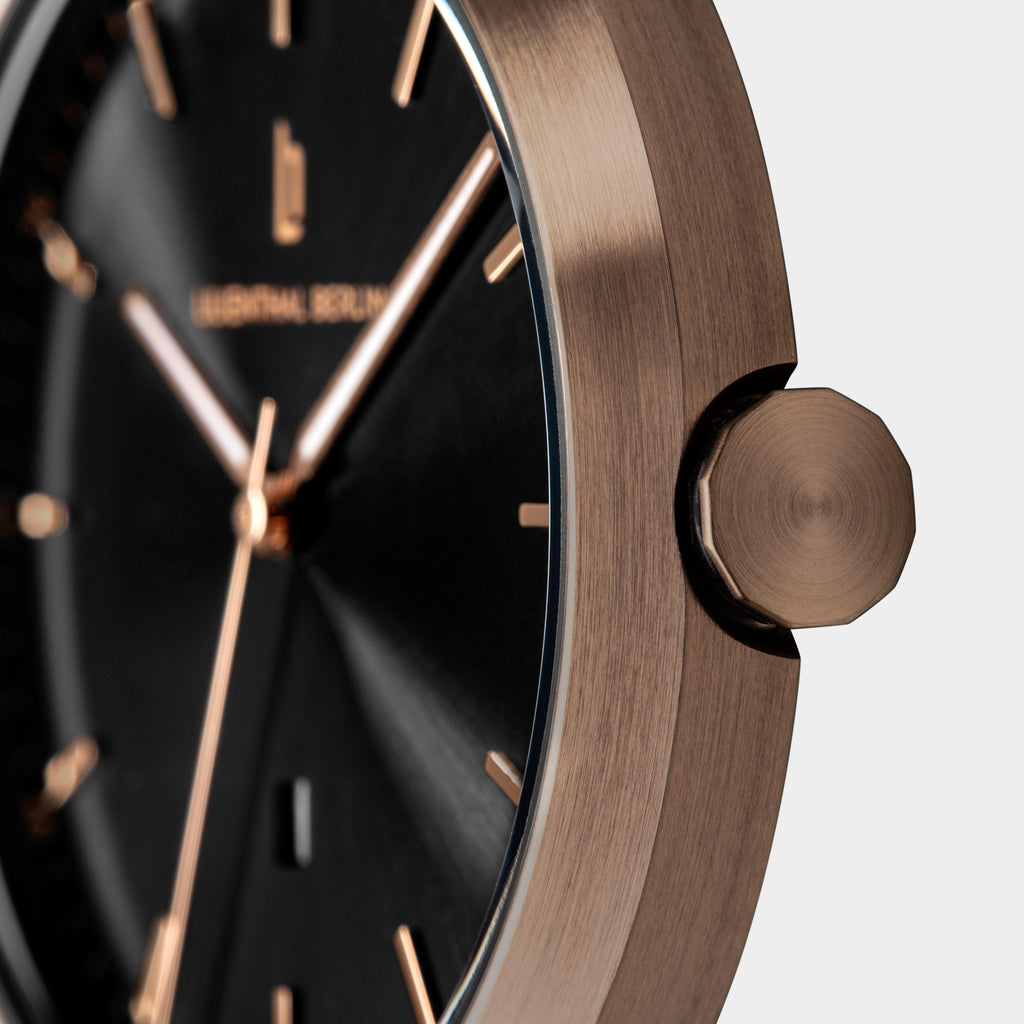 Close-up of a modern wristwatch with a brushed bronze case, black dial, and gold markers.