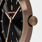 Close-up of a modern wristwatch with a brushed bronze case, black dial, and gold markers.
