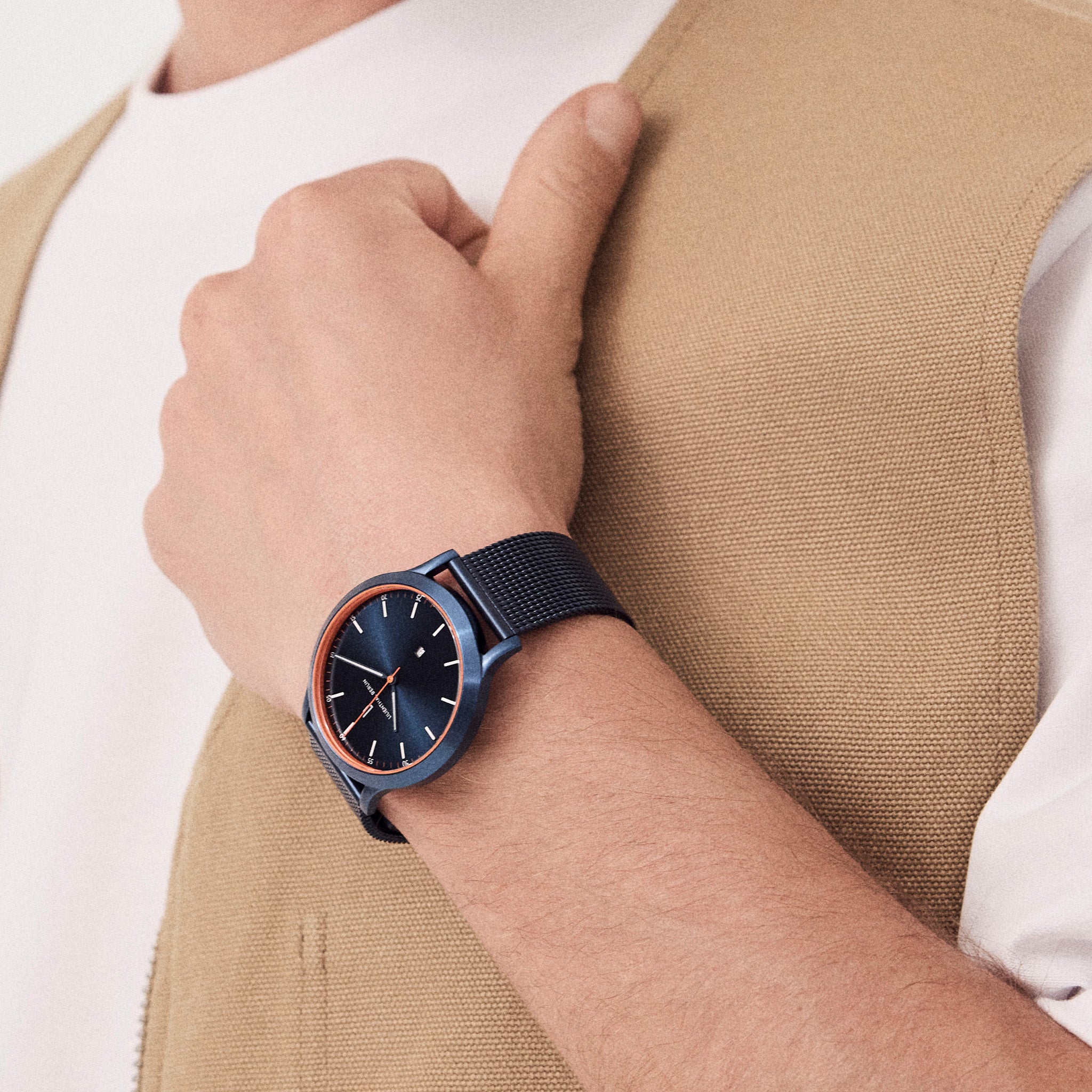 Elegant blue minimalist watch with mesh strap on wrist, paired with beige vest and white shirt