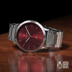 Stainless steel wristwatch with red dial and date display on wooden surface, German Design Award 2021 badge visible