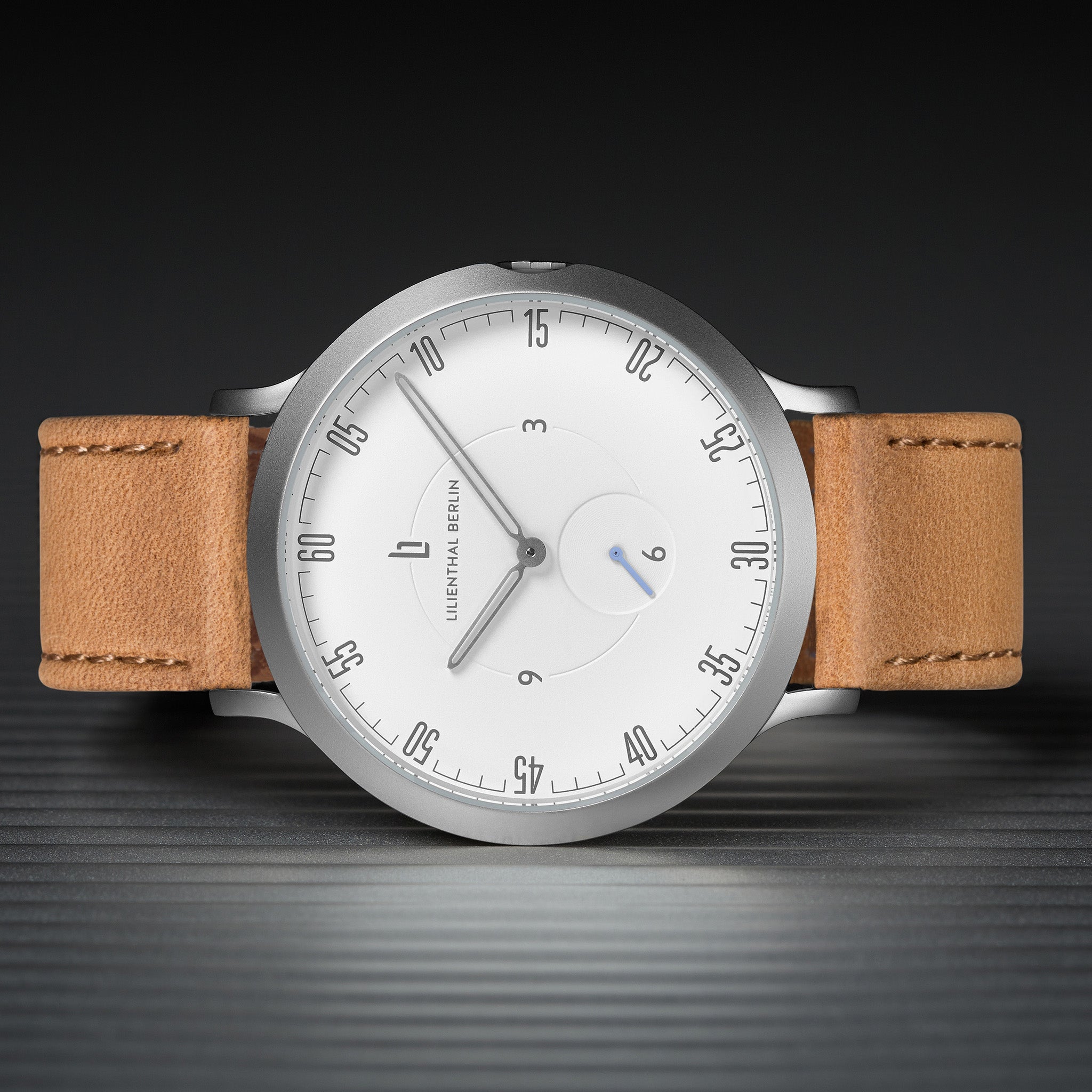 Minimalist silver watch with white dial and tan leather strap on dark textured surface