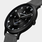 Minimalist black watch with black dial, white markers, and grey suede strap, close-up view.